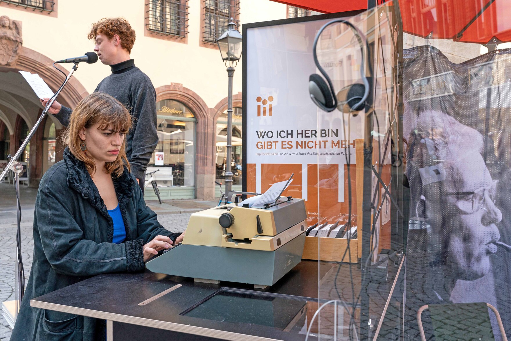 Eine Frau sitzt an einem Tisch vor einer Schreibmaschine, dahinter steht ein Mann an einem Mikrofon.